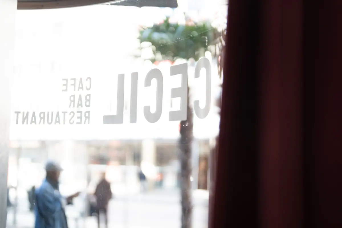 Blick von innen durch das Fenster mit dem Schriftzug «Cecil Café Bar Restaurant» auf den Platz